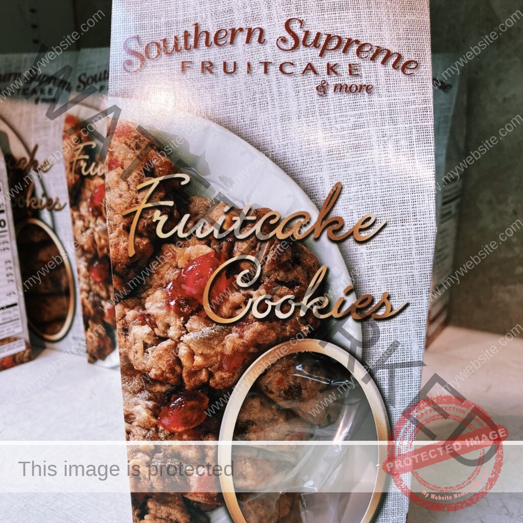 A tall white Southern Supreme gift box labeled "Fruitcake Cookies" in gold script beneath the "Southern Supreme Fruitcake & more" logo. The front shows a photo of cherry-and-nut-studded cookies and a clear oval window revealing the actual cookies inside. More boxes are lined up on the shelf behind.