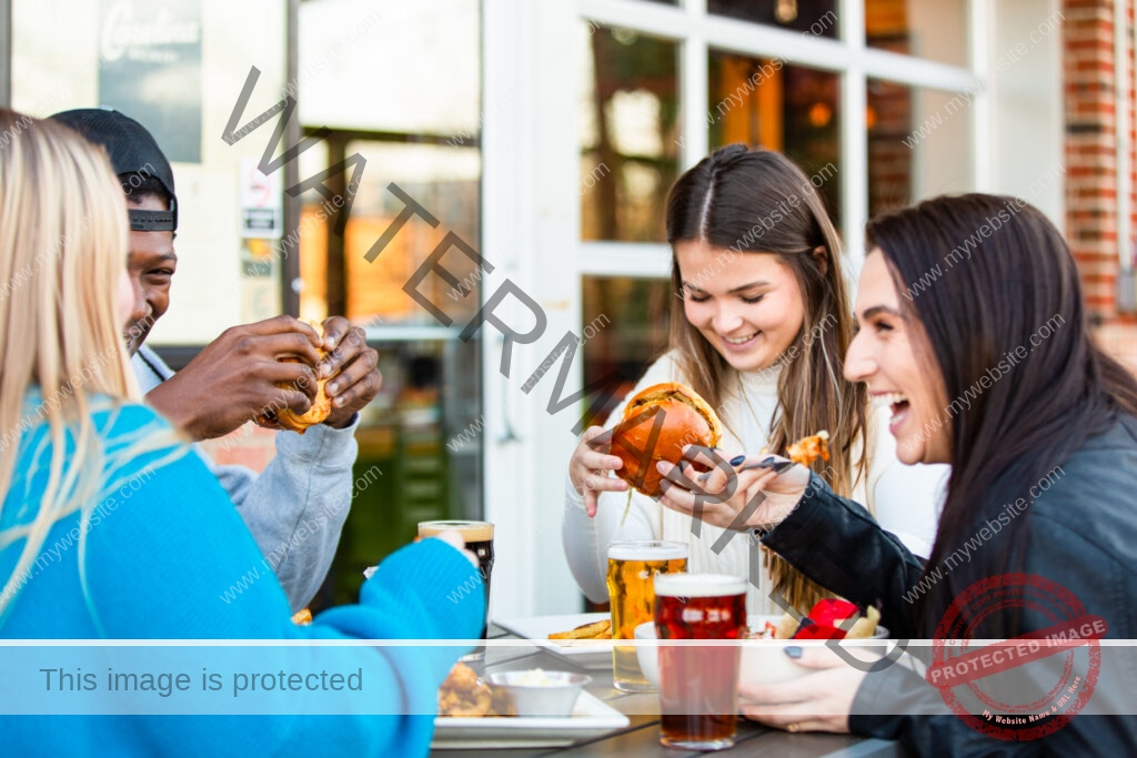 Four friends gathered around an outdoor table outside Carolina Brewery, eating burgers and laughing together. Glasses of light and dark beer sit on the table alongside plates of fries and shareable bowls. The brewery's windows and brick exterior, including a partial "Carolina" sign, are visible behind them at golden hour.