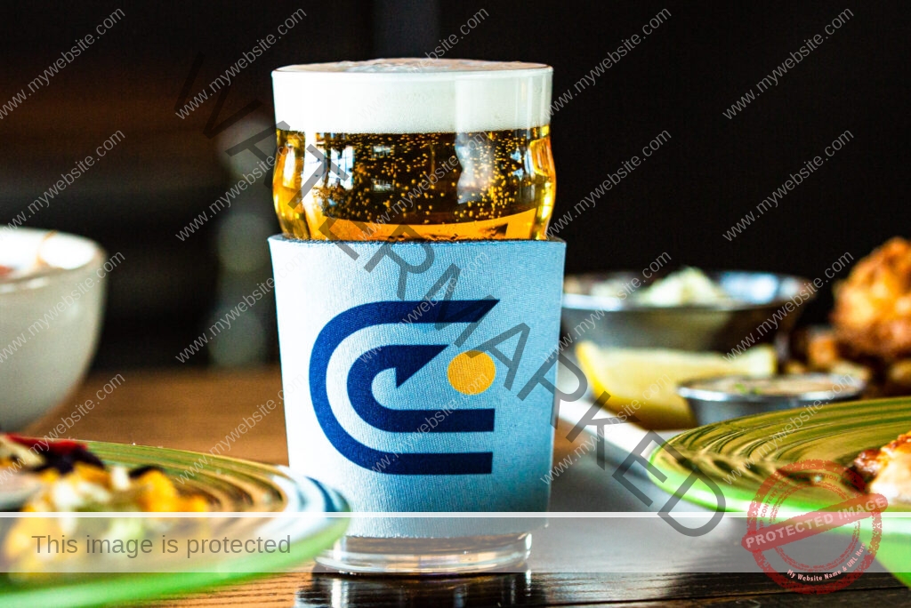 A full pint glass of golden, bubbly beer with a foamy head, wrapped in a light-blue koozie printed with Carolina Brewery's navy-and-gold "C" logo. The glass sits on a dark wooden table, framed by two green plates of food, a lemon wedge, and a ramekin in soft focus.