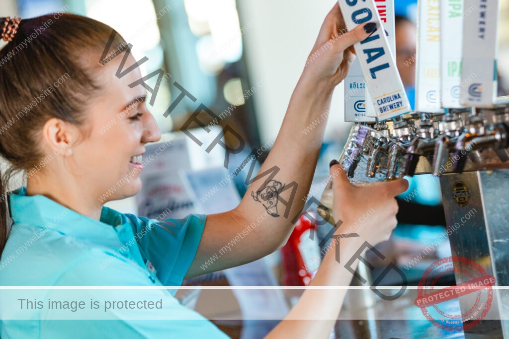 A profile view of a smiling bartender in a teal polo shirt pulling a Carolina Brewery tap handle to pour a draft beer. A row of branded tap handles labeled with beer styles (Kölsch, lager, IPA, Mexican) lines the bar, and a forearm tattoo is visible. The setting is a bright taproom bar.