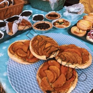 An outdoor bakery stand display at a farmers' market. In the foreground, several rustic open-faced tarts are topped with neatly fanned slices of roasted squash on flaky pastry crusts. Behind them, trays hold chocolate-glazed cakes in paper liners, small individual pies, a spinach quiche, and glazed pastries, all arranged on a turquoise daisy-print tablecloth.