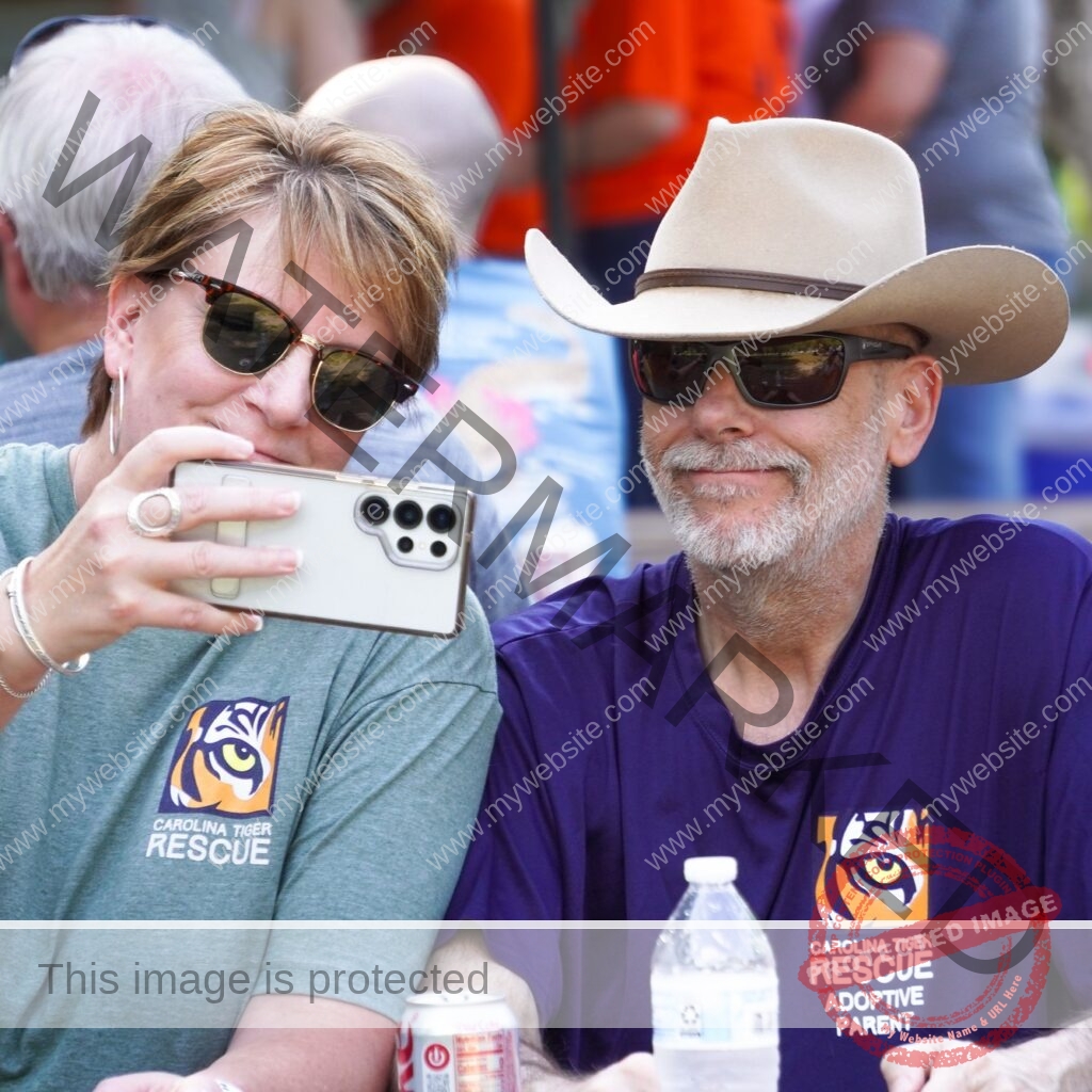 Two people sit close together at an outdoor event, both wearing Carolina Tiger Rescue T-shirts—one gray, one purple reading "Adoptive Parent." The person on the left, in sunglasses, holds up a phone to take a photo; the person on the right wears a tan cowboy hat and sunglasses and smiles toward the camera. Other attendees and a drink table are softly blurred in the background.
