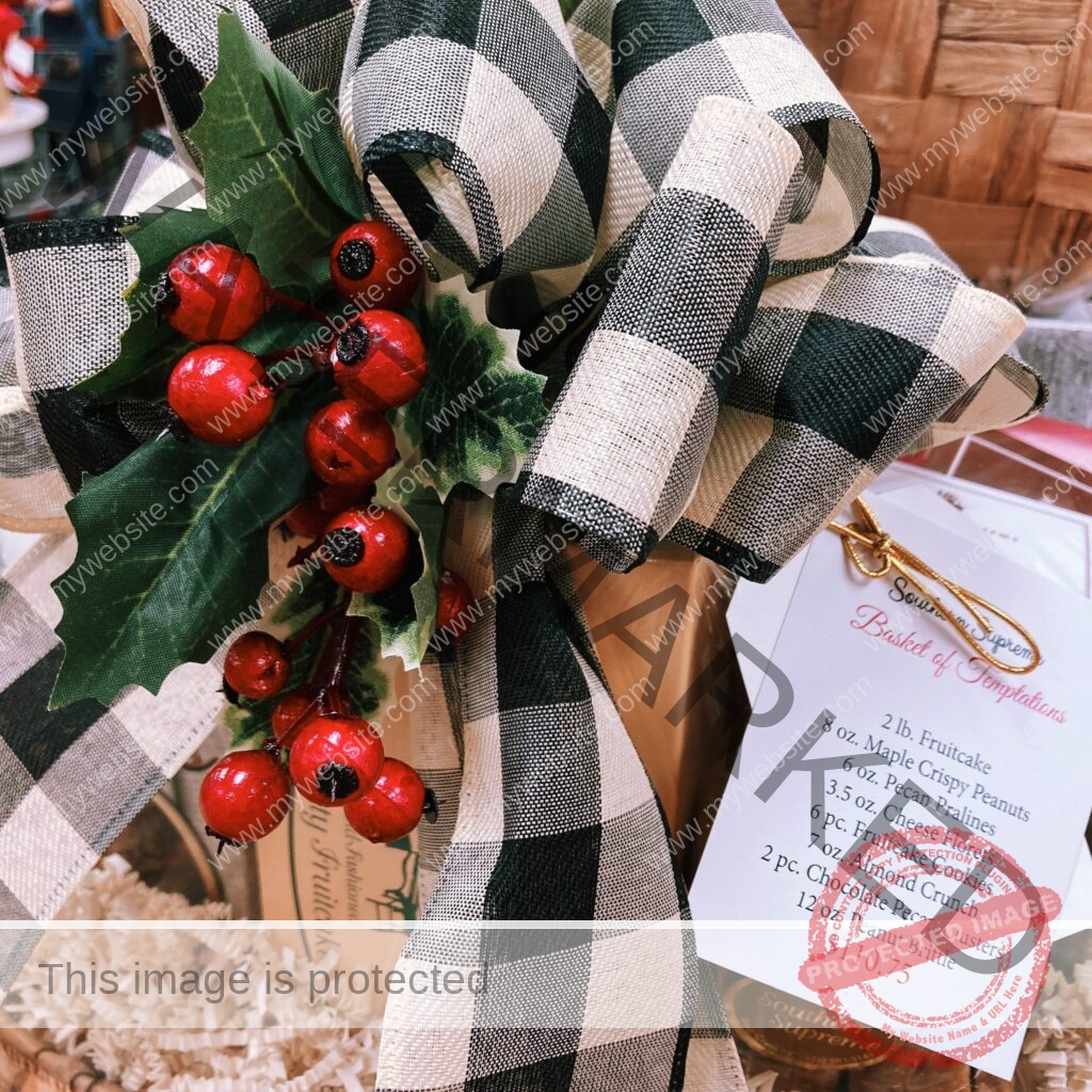 A close-up of a Southern Supreme gift basket tied with a large black-and-white buffalo-check bow accented with holly leaves and red berries. A boxed fruitcake sits inside amid paper shred. A white card labeled "Southern Supreme Basket of Temptations" lists the contents: 2 lb. fruitcake, 8 oz. maple crispy peanuts, 6 oz. pecan pralines, 6 pc. cheese florets, 6 pc. fruitcake cookies, 7 oz. almond crunch, 2 pc. chocolate pecan clusters, and 12 oz. peanut brittle, priced at $110.75.