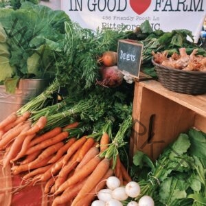 A farm stand display of freshly harvested root vegetables at the Fearrington Farmers' Market. Bundles of orange carrots with green tops, gold and red beets, and white salad turnips are arranged on a cloth-covered table beside a wooden crate, with leafy greens and a basket of shallots nearby. A small chalkboard reads "Beets," and a banner above reads "In Good Heart Farm" with a Pittsboro, NC address.