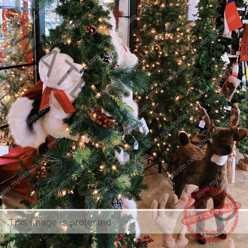Several fully lit and decorated artificial Christmas trees fill a showroom. A large white plush teddy bear in a red-and-white coat is nestled into the garland of one tree, and a life-size standing plush reindeer with antlers stands on the floor to the right. Trees are trimmed with warm lights, red berries, pinecones, and ornaments, with a North Carolina flag visible at upper right.