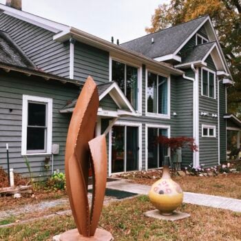 The Inn at Celebrity Dairy, a two-story gray clapboard farmhouse with white trim, large windows, and a covered entry marked 144. On the autumn lawn out front stand a tall abstract rusted-steel sculpture and a round yellow glazed-pottery vessel on a pedestal, with fall foliage and garden plantings around the house.