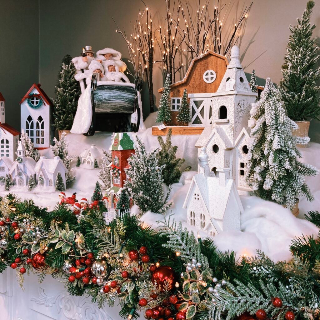 A detailed Christmas village arranged on white faux snow, with miniature white and red-roofed houses, steepled churches, and a wooden barn surrounded by frosted evergreen trees and twig lights. A Victorian-dressed family rides a sledge at the back, and small red-suited figures play in the snow. The foreground is framed by a lit garland of greenery, red berries, and silver and red ornaments.