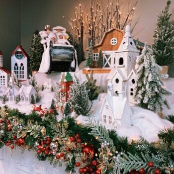 A detailed Christmas village arranged on white faux snow, with miniature white and red-roofed houses, steepled churches, and a wooden barn surrounded by frosted evergreen trees and twig lights. A Victorian-dressed family rides a sledge at the back, and small red-suited figures play in the snow. The foreground is framed by a lit garland of greenery, red berries, and silver and red ornaments.
