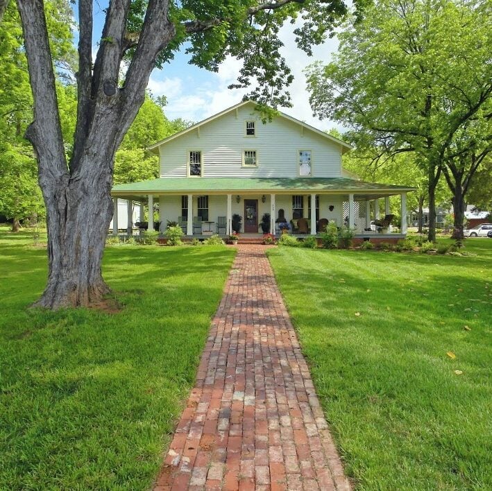 Exterior picture showing the front of 458 West Bed & Breakfast. A brick walkway leads to the charming two-story white craftsman home with a light green roof and wrap-around porch surrounded by green grass and large oak trees.