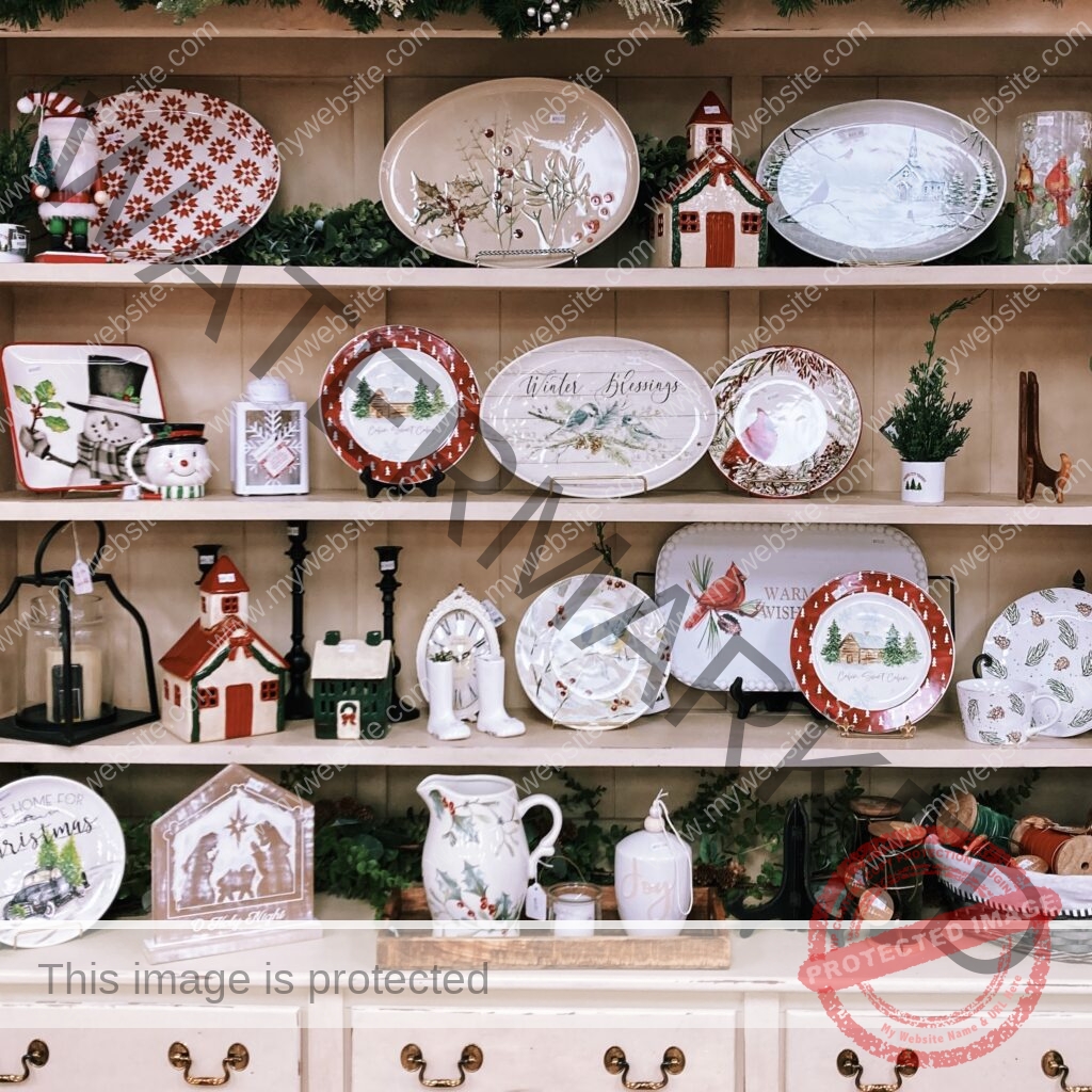 A cream-colored wooden hutch filled with holiday tableware and decor for sale. Shelves hold decorative plates and platters featuring poinsettias, holly, cardinals, snowmen, winter churches, and "Winter Blessings" and "Cabin Sweet Cabin" designs, alongside ceramic house figurines, candle lanterns, a holly-patterned pitcher, and a nativity "O Holy Night" plaque. Greenery garland trims the shelves.