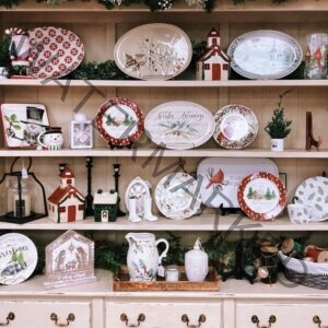 A cream-colored wooden hutch filled with holiday tableware and decor for sale. Shelves hold decorative plates and platters featuring poinsettias, holly, cardinals, snowmen, winter churches, and "Winter Blessings" and "Cabin Sweet Cabin" designs, alongside ceramic house figurines, candle lanterns, a holly-patterned pitcher, and a nativity "O Holy Night" plaque. Greenery garland trims the shelves.