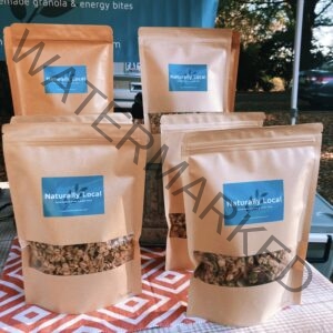 Five kraft-paper stand-up pouches of "Naturally Local" homemade granola arranged on a patterned cloth at the Fearrington Farmers' Market. Each bag has a blue label reading "Naturally Local – homemade granola & power bites" and a clear window showing the oat-and-seed granola inside, with two marked "Pecan." A blue vendor banner reading "granola & energy bites" hangs behind the display, with trees and parked vehicles beyond the tent.