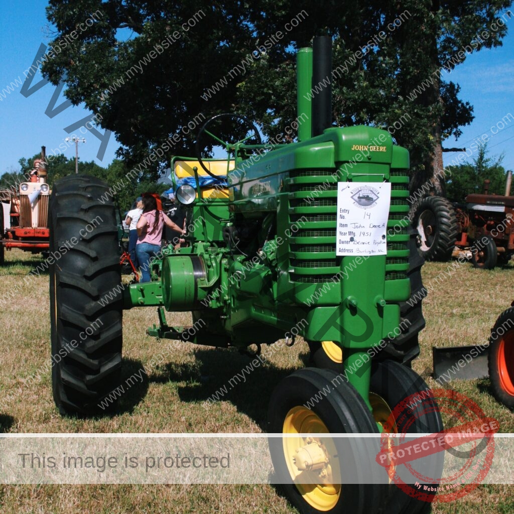 A fully restored green-and-yellow John Deere Model A tractor stands on grass at an outdoor farm heritage show. A handwritten exhibitor card hangs on its grille listing it as Entry No. 14, a John Deere A manufactured in 1951. Other antique tractors in red and cream are lined up on either side, with attendees browsing in the background beneath large shade trees and a clear blue sky.