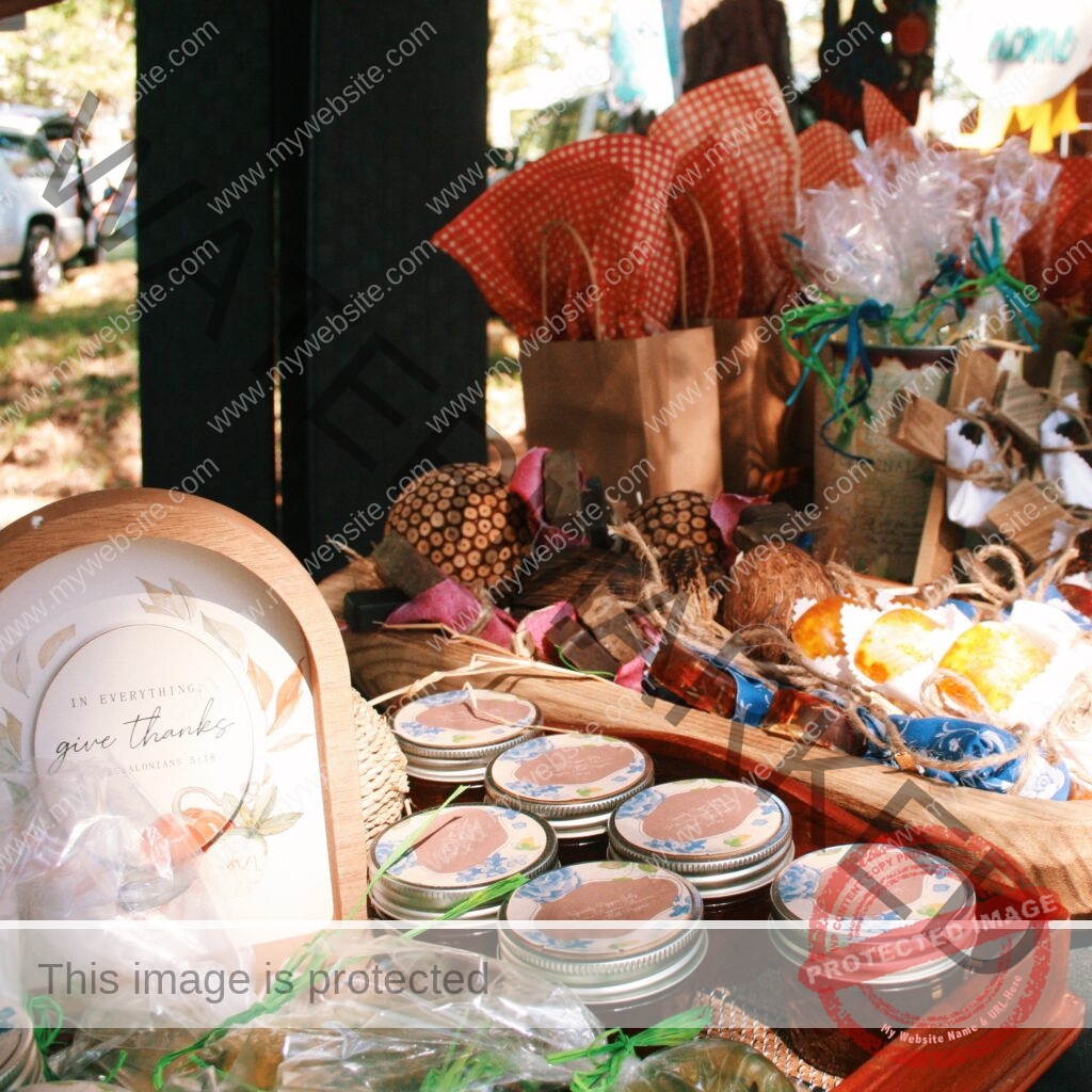 A sunlit vendor table at an outdoor market displays an array of handmade goods: rows of small mason jars of homemade jelly with decorative floral lids arranged in a wooden trencadis tray, clear-wrapped baked treats tied with green ribbon, and rustic gift items including soaps and twine-tied bundles. A small wooden-framed sign on the left reads "In everything, give thanks." A brown paper gift bag with red gingham tissue sits in the background.
