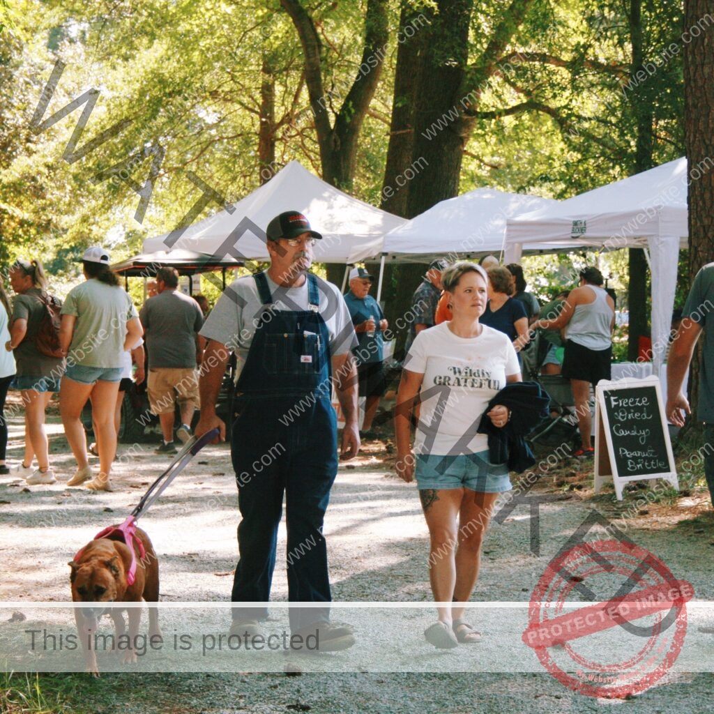 A man in denim overalls and a woman in a white "Wildly Grateful" T-shirt and denim shorts walk a leashed brown dog along a gravel path at an outdoor festival on a sunny day. White pop-up vendor tents line the path beneath tall shade trees, with other attendees browsing. A small chalkboard sign at right advertises "Freeze Dried Candy" and "Peanut Brittle."