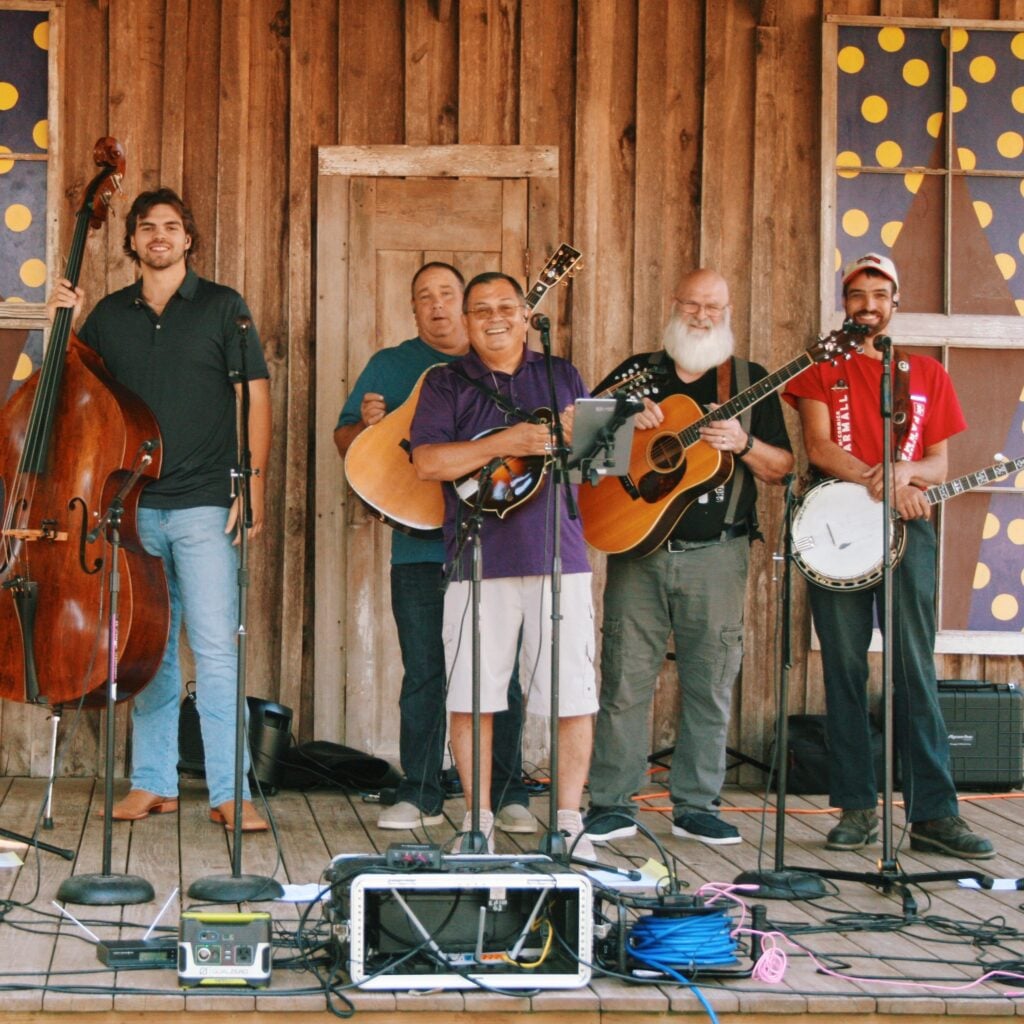 Six smiling musicians stand in a row on a weathered wooden stage, posing with their bluegrass instruments before or after a performance. From left to right, they hold a resonator guitar (Dobro), an upright bass, a mandolin, an acoustic guitar, and a banjo, with microphones on stands in front of each player. The backdrop is a rustic barn-wood wall with an old paneled door and two windows decorated with whimsical purple-and-yellow polka-dot designs. Sound equipment, cables, and instrument cases are arranged across the stage floor in the foreground.
