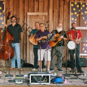Six smiling musicians stand in a row on a weathered wooden stage, posing with their bluegrass instruments before or after a performance. From left to right, they hold a resonator guitar (Dobro), an upright bass, a mandolin, an acoustic guitar, and a banjo, with microphones on stands in front of each player. The backdrop is a rustic barn-wood wall with an old paneled door and two windows decorated with whimsical purple-and-yellow polka-dot designs. Sound equipment, cables, and instrument cases are arranged across the stage floor in the foreground.