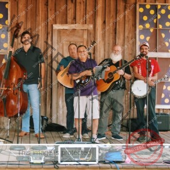 Six smiling musicians stand in a row on a weathered wooden stage, posing with their bluegrass instruments before or after a performance. From left to right, they hold a resonator guitar (Dobro), an upright bass, a mandolin, an acoustic guitar, and a banjo, with microphones on stands in front of each player. The backdrop is a rustic barn-wood wall with an old paneled door and two windows decorated with whimsical purple-and-yellow polka-dot designs. Sound equipment, cables, and instrument cases are arranged across the stage floor in the foreground.