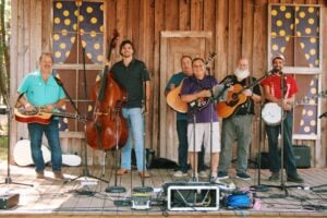 Six smiling musicians stand in a row on a weathered wooden stage, posing with their bluegrass instruments before or after a performance. From left to right, they hold a resonator guitar (Dobro), an upright bass, a mandolin, an acoustic guitar, and a banjo, with microphones on stands in front of each player. The backdrop is a rustic barn-wood wall with an old paneled door and two windows decorated with whimsical purple-and-yellow polka-dot designs. Sound equipment, cables, and instrument cases are arranged across the stage floor in the foreground.
