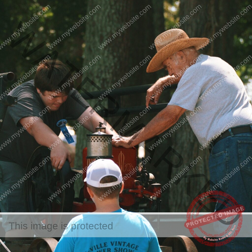 Under shade trees at an outdoor antique-power show, an older man in a straw hat leans over a small restored vintage engine, resting his hand on it while talking with a teenager who bends in to look closely. A blue prize ribbon is pinned to the teen's shirt. A younger child in a backwards white cap and blue T-shirt watches from the foreground. The engine is mounted on a wheeled cart, and trees fill the background.