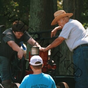 Under shade trees at an outdoor antique-power show, an older man in a straw hat leans over a small restored vintage engine, resting his hand on it while talking with a teenager who bends in to look closely. A blue prize ribbon is pinned to the teen's shirt. A younger child in a backwards white cap and blue T-shirt watches from the foreground. The engine is mounted on a wheeled cart, and trees fill the background.