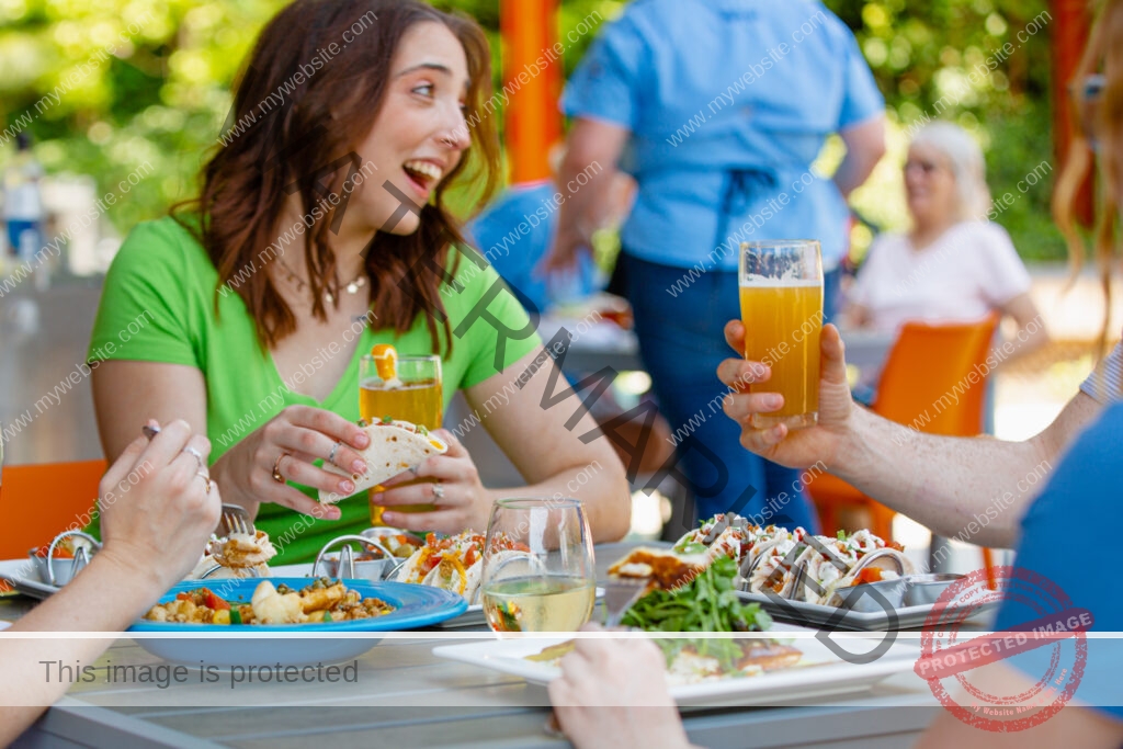 A group of friends dine at an outdoor table on a sunny patio, sharing plates of tacos and bowls of food. A smiling woman in a green shirt holds a taco and a beer; a hand at right raises a glass of hazy beer in a toast. A server and other diners are visible in the background among green trees and orange patio furniture.