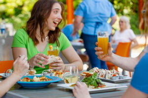 A group of friends dine at an outdoor table on a sunny patio, sharing plates of tacos and bowls of food. A smiling woman in a green shirt holds a taco and a beer; a hand at right raises a glass of hazy beer in a toast. A server and other diners are visible in the background among green trees and orange patio furniture.