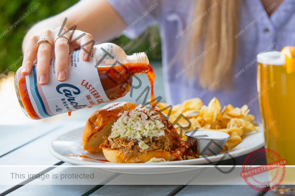A close-up of a person pouring Carolina Brewery's Eastern Carolina BBQ Sauce from a glass bottle over a pulled-pork sandwich topped with coleslaw on a toasted bun. The plate includes kettle chips and a ramekin of dressing, with a glass of beer beside it on an outdoor table.