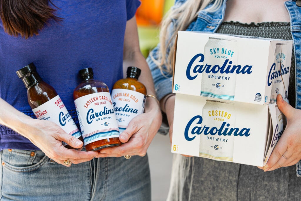 A close-up of two people holding Carolina Brewery products. One holds three glass bottles labeled Sriracha Steak Sauce, Eastern Carolina BBQ Sauce, and Carolina Gold BBQ Sauce; the other holds stacked six-pack cartons of Sky Blue Kölsch and Costero Lager cans. The packaging features the brewery's blue script logo and wave motif.