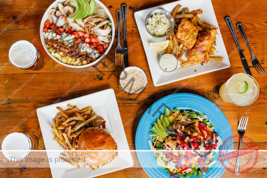 An overhead view of a wooden table set with several dishes and drinks. The spread includes a Cobb salad with grilled chicken, tomato, bacon, blue cheese, and avocado; a plate of fish and chips with slaw and tartar sauce; a burger with hand-cut fries; and a Southwest-style salad with fried chicken, avocado, cheese, and tortilla strips on a blue plate. Three glasses of beer and a lime-garnished margarita are arranged around the plates.