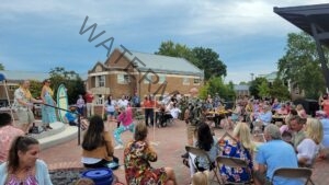 A festive outdoor gathering on a brick patio, where a crowd in Hawaiian shirts, floral dresses, and flower leis watches a limbo contest. A participant in colorful patterned leggings bends backward to pass under a wooden limbo bar held by two people, while seated guests at long tables look on and cheer. A decorative surfboard reading "El Mirage" stands on a small stage at left, and a brick building with a metal roof and mature trees rise behind the party under a partly cloudy sky.