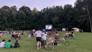 An outdoor movie night on a grassy field at dusk. Families and children sit on folding camp chairs, blankets, and the grass, facing a large inflatable projection screen showing an animated film. A person in a white "Supervisor" shirt stands among the crowd, and a vendor tent and trailer are set up to the right near soccer goals at the field's edge. Tall green trees line the background under a soft lavender evening sky.