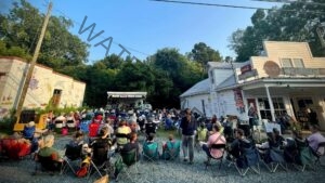 A crowd of dozens sits in folding camp chairs on a gravel lot, facing a small covered stage where a band performs for the Bynum Front Porch music series. The white clapboard Bynum General Store stands at right with vintage Coca-Cola signs, a covered porch, and a free book exchange visible through the doorway. A building painted with a colorful animal mural is at left, framed by tall green trees under a clear blue evening sky.