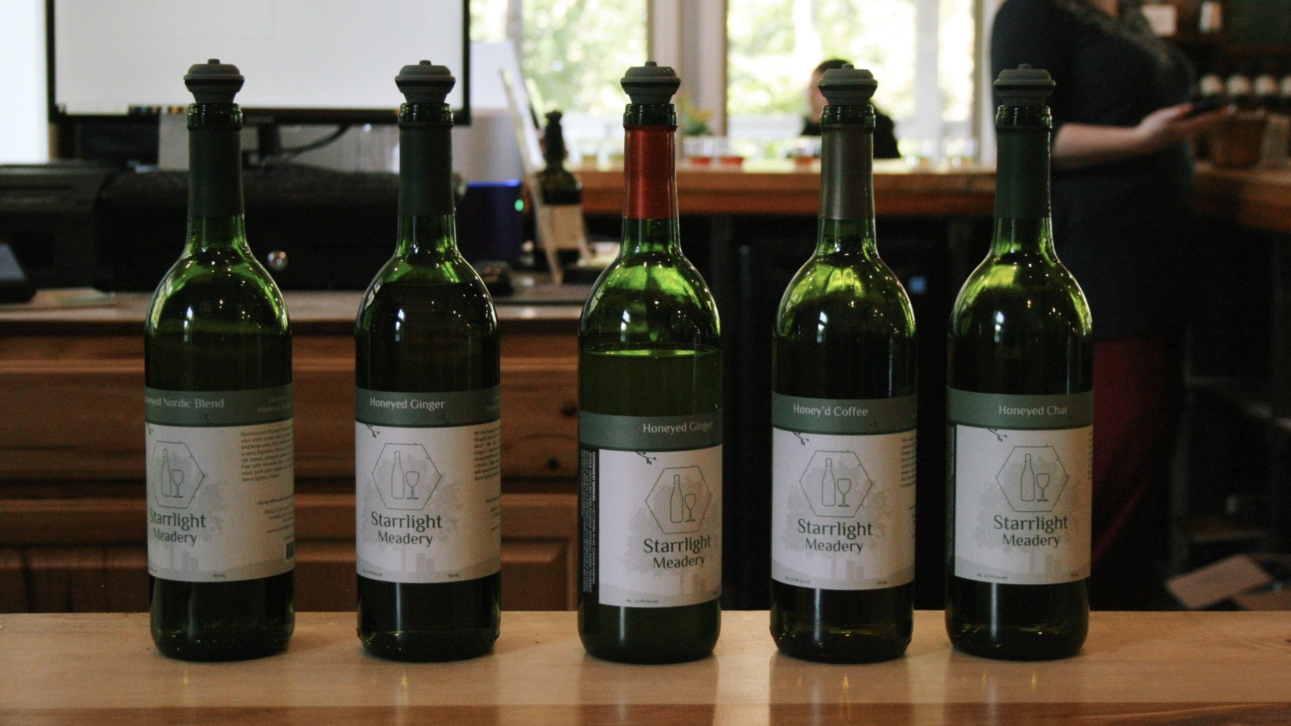 Five green glass bottles of Starrlight Meadery mead stand in a row on a wood tasting-room bar, each with a cream label bearing the meadery's hexagon bottle-and-glass logo. Visible varieties include Honeyed Nordic Blend, Honeyed Ginger, Honey'd Coffee, and Honeyed Chai. Behind the bottles, a staff member stands at the bar near windows and shelves of products.
