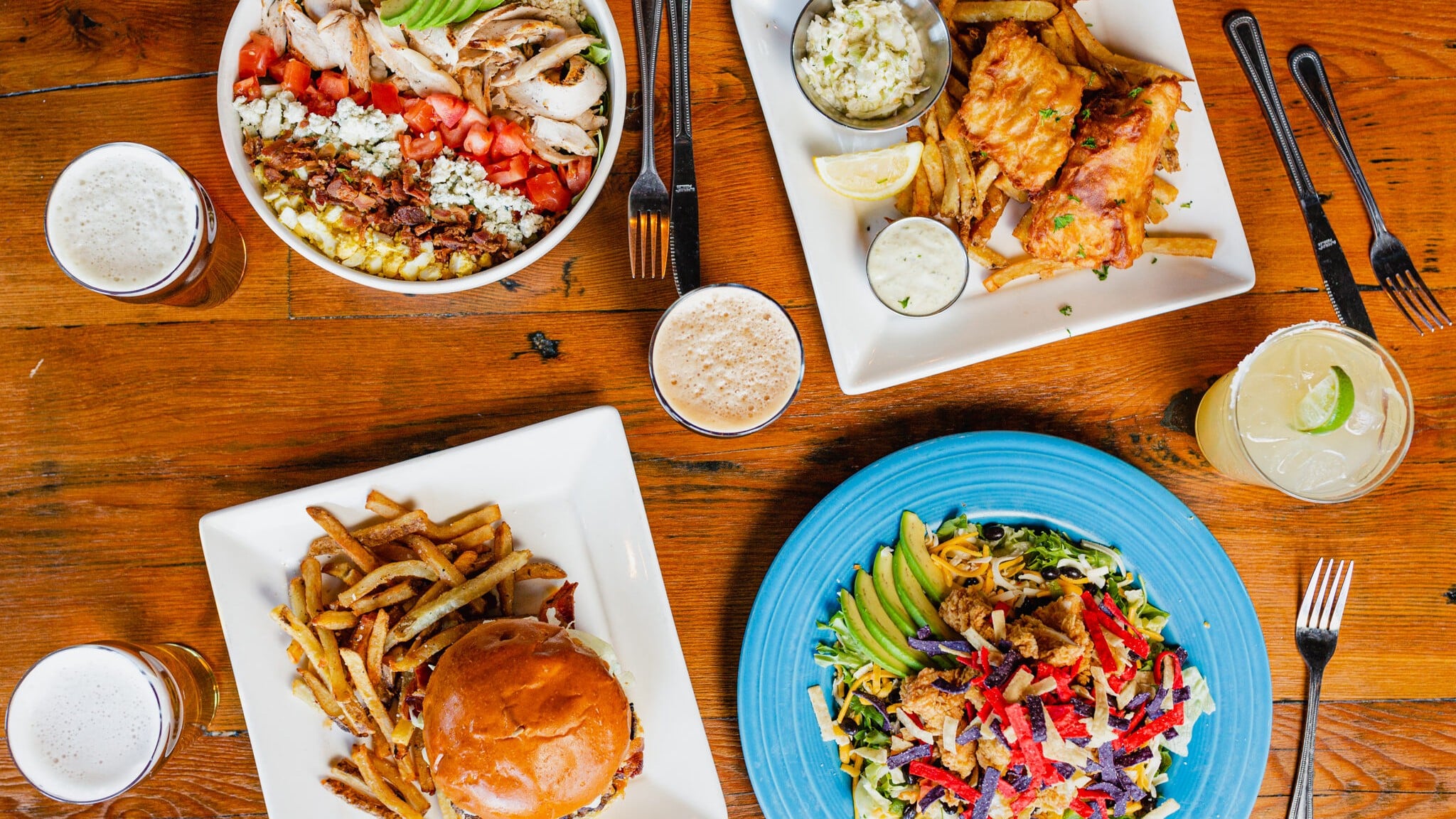 An overhead view of a wooden table set with several dishes and drinks. The spread includes a Cobb salad with grilled chicken, tomato, bacon, blue cheese, and avocado; a plate of fish and chips with slaw and tartar sauce; a burger with hand-cut fries; and a Southwest-style salad with fried chicken, avocado, cheese, and tortilla strips on a blue plate. Three glasses of beer and a lime-garnished margarita are arranged around the plates.
