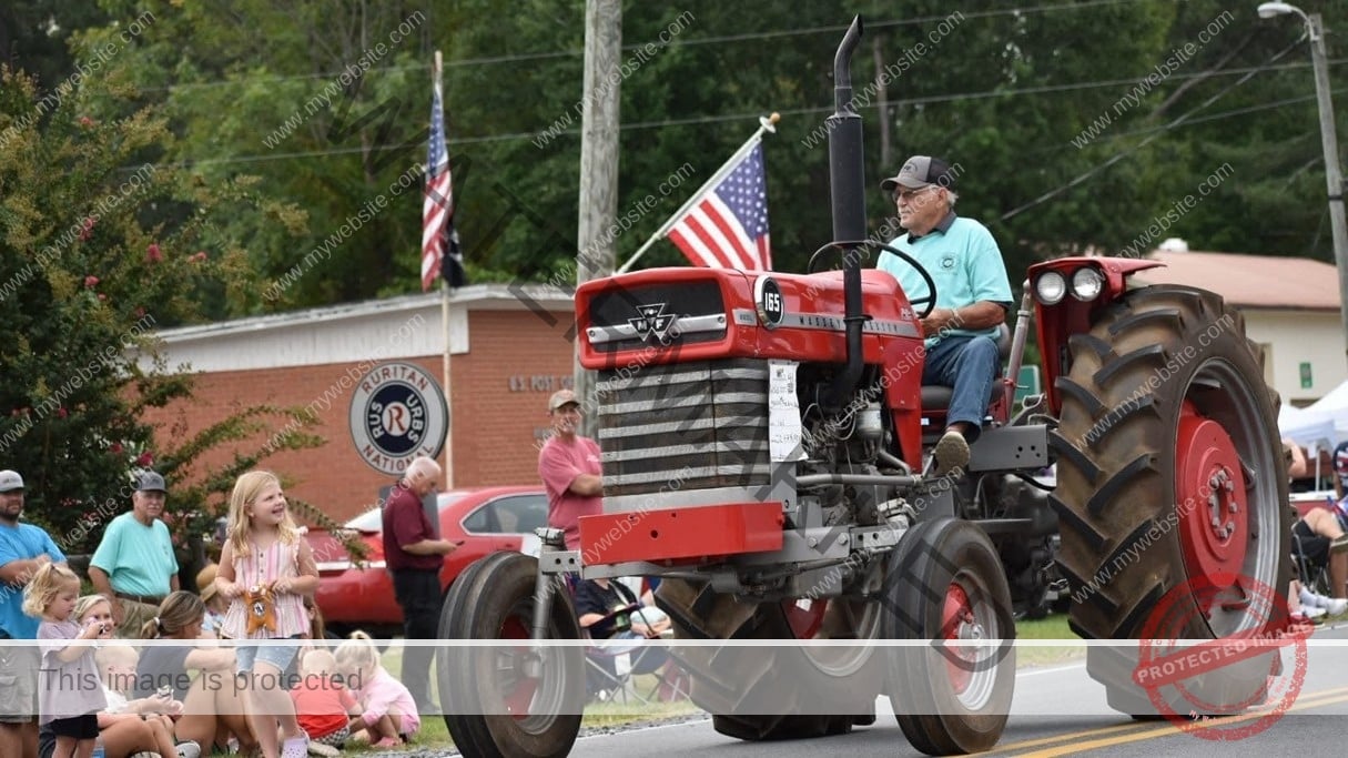A man in a teal shirt and ball cap drives a restored vintage red Massey Ferguson 165 tractor down a road during a small-town parade. American flags fly from the tractor, and spectators—including young children seated on the grass—watch from the roadside on a partly cloudy day. Behind them stands a brick building with a Ruritan National emblem and a U.S. Post Office sign, framed by green trees and crepe myrtle in bloom.