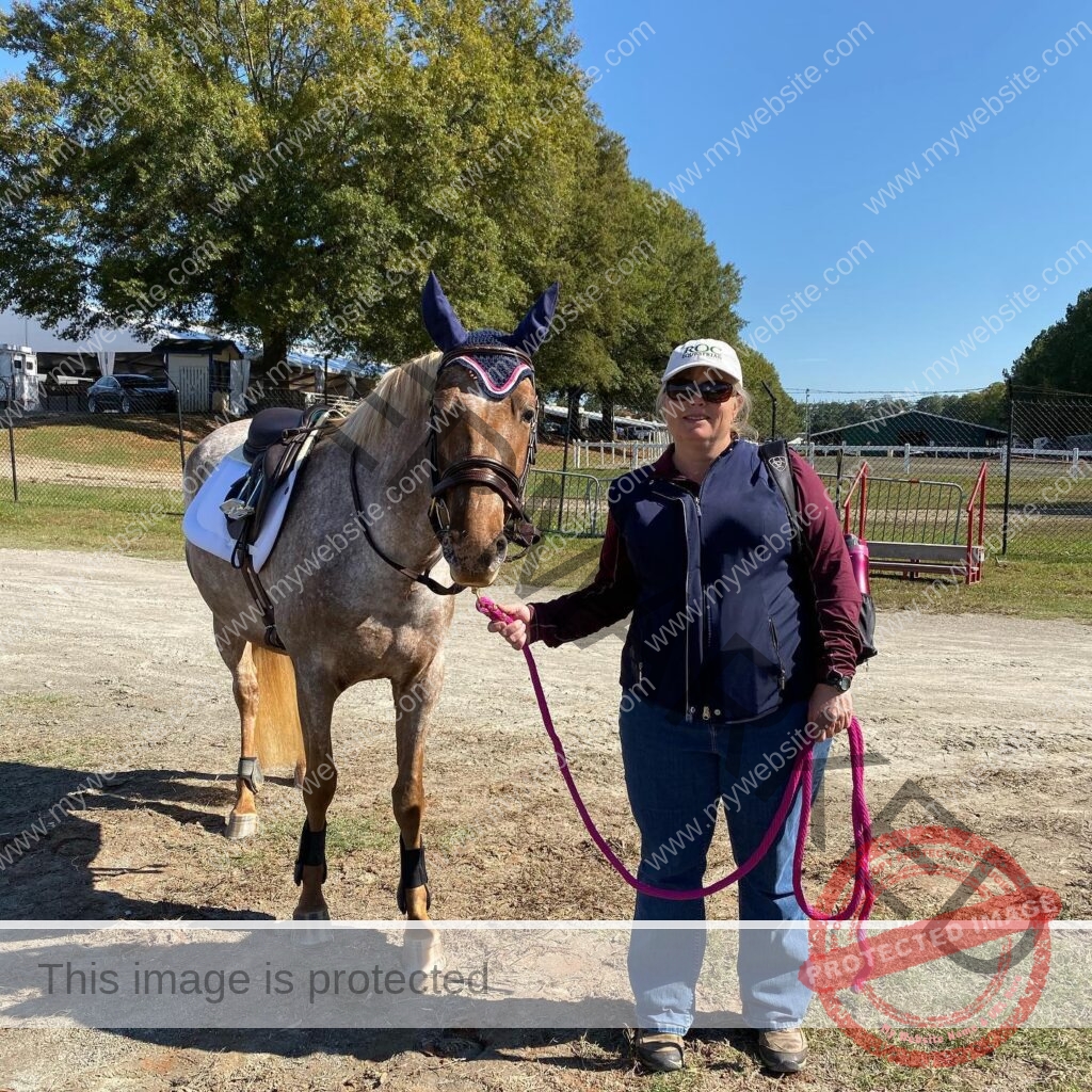 A woman in a white ROC Equestrian cap, sunglasses, and a maroon-and-navy jacket stands smiling beside a saddled appaloosa pony wearing a navy ear bonnet and bridle. She holds a bright pink lead rope. Behind them: a trailer parking area, jump standards, and trees under a clear blue sky.