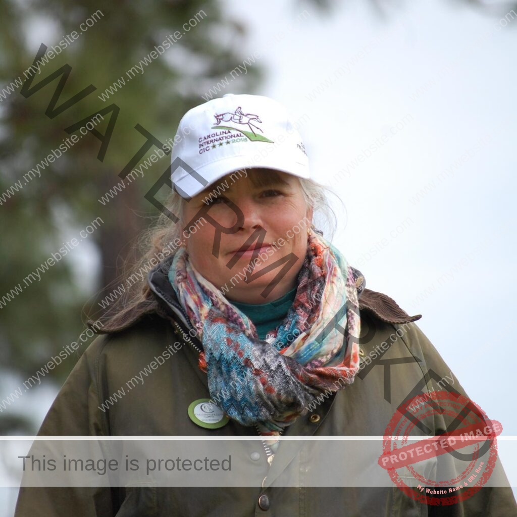 Close-up portrait of a woman with light blonde hair smiling softly at the camera. She wears a white Carolina International CIC 2018 ball cap, a brightly patterned woven scarf in teal, orange, and pink, and an olive-green waxed canvas jacket with a Carolina Club member button on the lapel. A pine tree and overcast sky blur behind her.