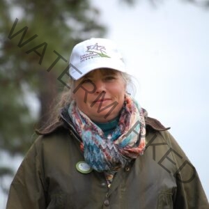 Close-up portrait of a woman with light blonde hair smiling softly at the camera. She wears a white Carolina International CIC 2018 ball cap, a brightly patterned woven scarf in teal, orange, and pink, and an olive-green waxed canvas jacket with a Carolina Club member button on the lapel. A pine tree and overcast sky blur behind her.