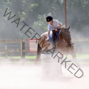 A young rider in a black helmet and white t-shirt leans forward in the saddle, gripping the reins as a roan horse pivots tightly around a white barrel. Dust kicks up around the horse's legs in the warm sunlight. A wooden fence and trees frame the riding ring in the background.