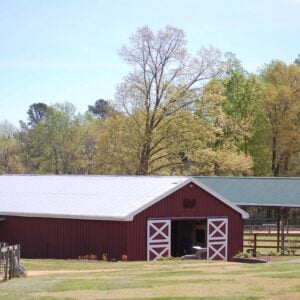 Exterior view of a tidy red barn with white cross-buck sliding doors and a silver metal roof at New Beginnings Farm. A green-roofed covered arena extends to the right, and post-and-rail fencing borders a grassy paddock and gravel drive in the foreground. Mature hardwood trees in bright spring green surround the property under a clear blue sky.