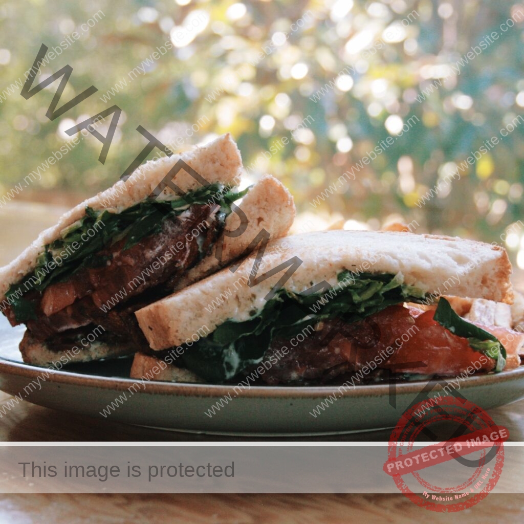 A halved sandwich stacked on a speckled stoneware plate, set on a wood table outdoors. The sandwich is made on thick white bread and layered with crisp bacon, ripe red tomato slices, fresh leafy greens, and a creamy pesto-flecked mayonnaise. Dappled sunlight filters through blurred green and golden trees in the background, giving the scene a warm, late-afternoon glow.