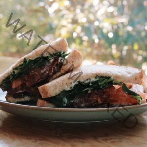 A halved sandwich stacked on a speckled stoneware plate, set on a wood table outdoors. The sandwich is made on thick white bread and layered with crisp bacon, ripe red tomato slices, fresh leafy greens, and a creamy pesto-flecked mayonnaise. Dappled sunlight filters through blurred green and golden trees in the background, giving the scene a warm, late-afternoon glow.
