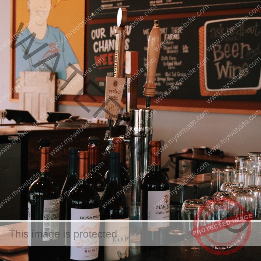 A close-up of the bar area at Cafe Root Cellar. A polished stainless beer-tap tower with wooden tap handles—one labeled "volio" sparkling white wine—rises at center. In front of it stand several bottles of wine, including Doña Paula Estate Malbec, a Pinot Noir, and a Marqués de Casa Concha Carménère, with copper and dark foil tops. Rows of clean, upturned drinking glasses line the dark countertop to the right. Behind the bar, the menu board and the edge of the Chef Cuni pop-art mural are visible, including a chalkboard reading "Ask us about our Beer and Wine."