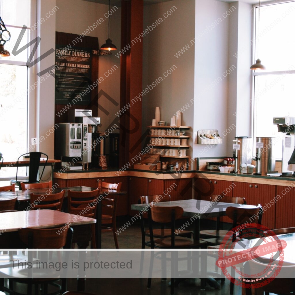 A warmly lit café interior with polished concrete floors and an assortment of wood tables and bentwood-style chairs. Along the back wall, a self-serve beverage station holds coffee brewers, stacked mugs on wooden shelving, and stainless iced-tea dispensers labeled "sweet" and "unsweet," set on light wood counters with rust-red cabinetry below. A large dark sign on the wall reads "The Root Cellar Family Dinners" and "Paleo Family Dinners," detailing the take-home meal program. Tall windows on either side let in daylight, and industrial pendant lights hang from the ceiling.