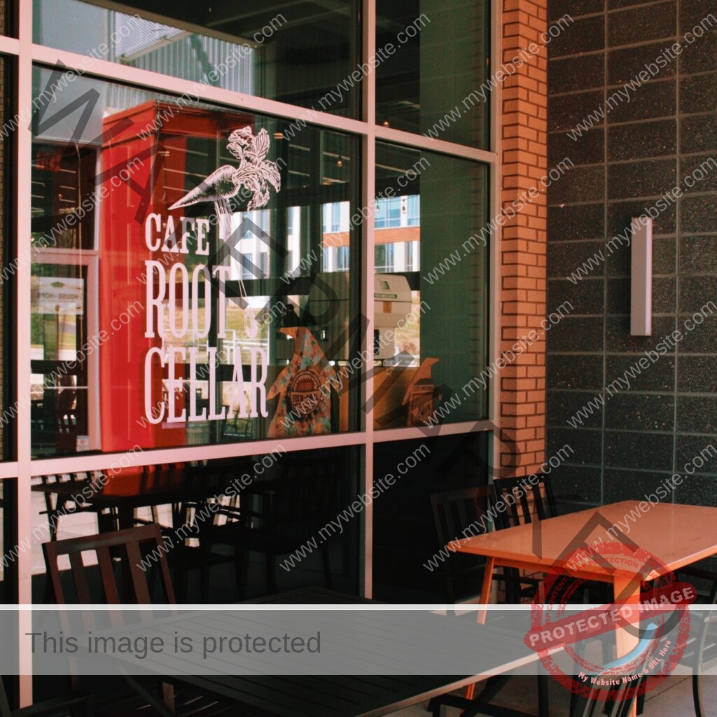 The storefront of Cafe Root Cellar viewed from the covered outdoor patio. A large plate-glass window bears the café's name in white lettering alongside its logo—a stylized heron perched on a fork with leafy plumage. Through the glass, a red refrigerator and decorative penguin figures (including one labeled "Feed-Well Fridge") are visible inside. The building combines red brick and dark speckled tile, and metal patio tables with chairs sit on the concrete in the warm, low-angled light.