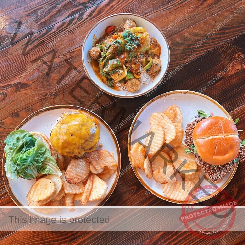 An overhead view of three plates arranged on a rich, dark wood table. At top is a white bowl holding a yellow curry over white rice, with meatballs, peppers, and fresh herbs. At bottom left, a white plate holds a cheeseburger topped with melted cheddar on a toasted bun, served with butter lettuce and a side of crinkle-cut waffle fries. At bottom right, a white plate holds a sandwich with a nut- or seed-crusted patty and microgreens on a glossy brioche bun, also with waffle fries. All three plates have warm, rust-toned rims.