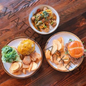 An overhead view of three plates arranged on a rich, dark wood table. At top is a white bowl holding a yellow curry over white rice, with meatballs, peppers, and fresh herbs. At bottom left, a white plate holds a cheeseburger topped with melted cheddar on a toasted bun, served with butter lettuce and a side of crinkle-cut waffle fries. At bottom right, a white plate holds a sandwich with a nut- or seed-crusted patty and microgreens on a glossy brioche bun, also with waffle fries. All three plates have warm, rust-toned rims.