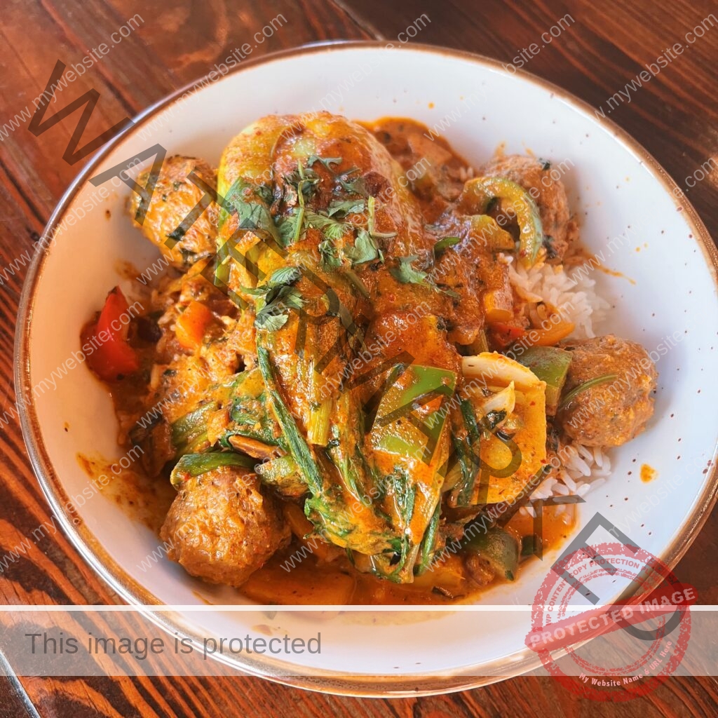 A close-up overhead shot of a white, rust-rimmed bowl filled with a vibrant yellow-orange curry served over white rice. The dish features tender pork meatballs, sliced green and red bell peppers, leafy greens, and chunks of vegetables in a rich, creamy sauce, finished with a scatter of fresh cilantro. The bowl rests on a dark, warm-toned wood table.