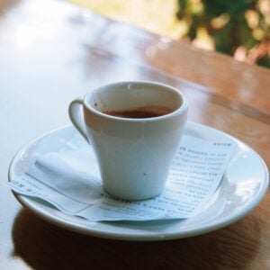 A small white ceramic cup holding a dark espresso sits on a matching white saucer, photographed in warm natural light. The cup and saucer rest on a folded printed paper menu headed "Cafe Root Cellar Dinner," with menu items partially legible beneath. The table has a glossy wood-toned surface, and softly blurred green and golden foliage is visible through a window in the background.